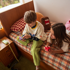 Two children sitting on a wooden bed with a plaid sheet, making a call on the Waffle Home